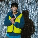 Lee Jae-wook memegang mikrofon dengan rompi kuning di depan tirai gemerlap dalam serial Dead-End Job
