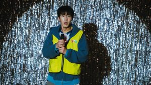 Lee Jae-wook memegang mikrofon dengan rompi kuning di depan tirai gemerlap dalam serial Dead-End Job