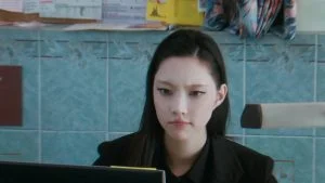 Member ILLIT berjas hitam duduk di depan monitor dalam teaser You're Under Arrest