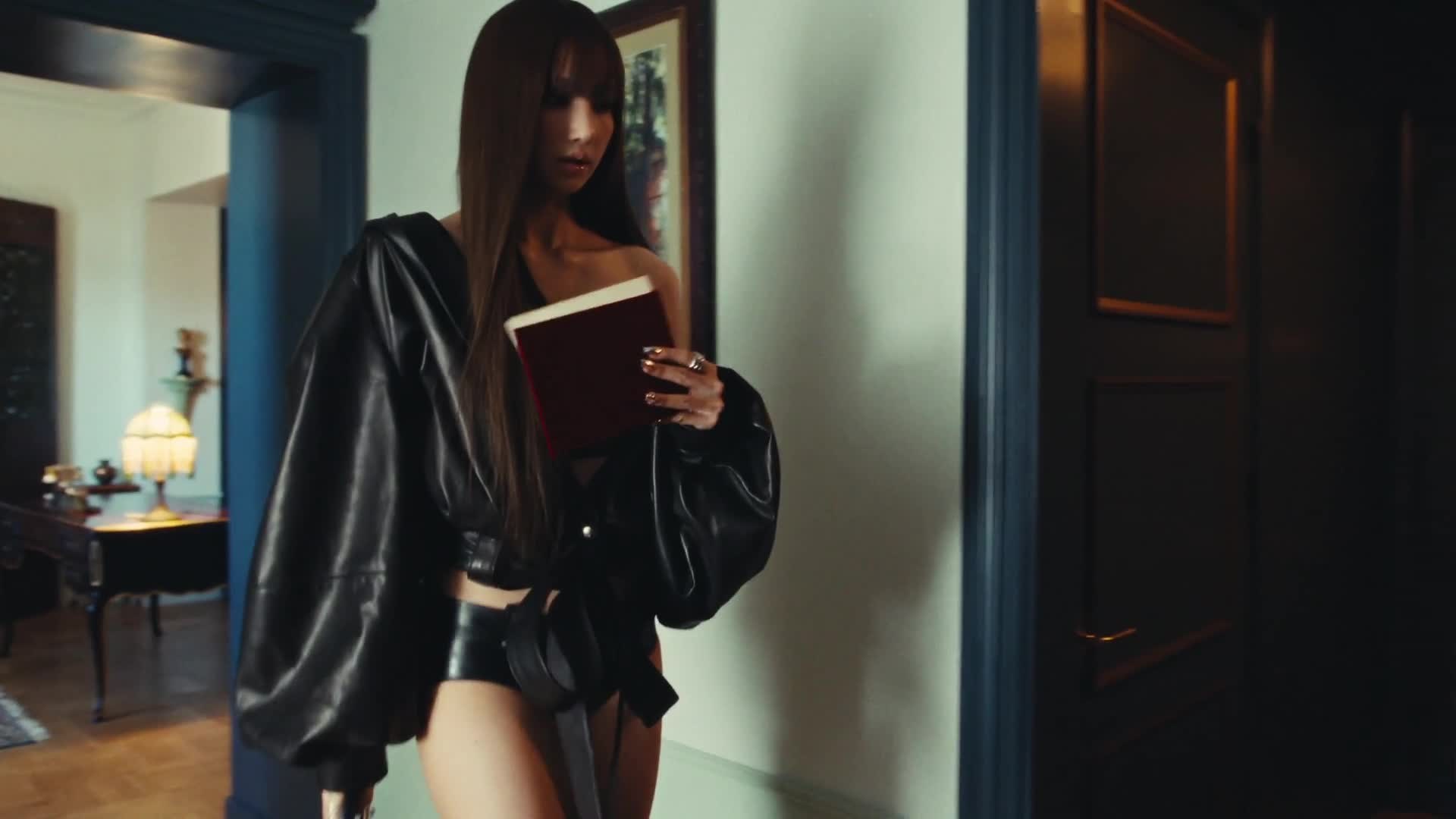 LISA berjaket kulit hitam berjalan sambil membawa buku bersampul merah dalam teaser MV Tarantino