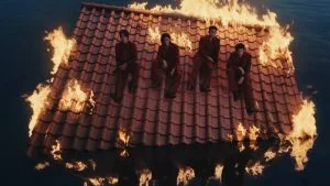 Keempat member LNGSHOT duduk di atap merah yang terbakar di tengah air dalam MV Masterpiece