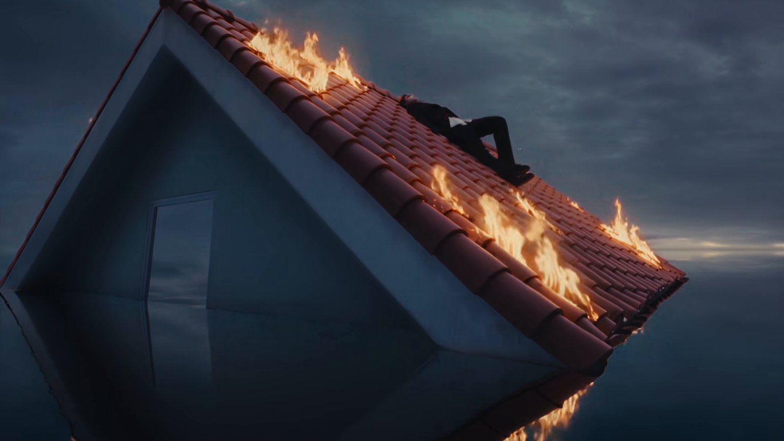 Atap rumah merah terbakar di tengah air dengan seorang member LNGSHOT berbaring di atasnya dalam MV Masterpiece
