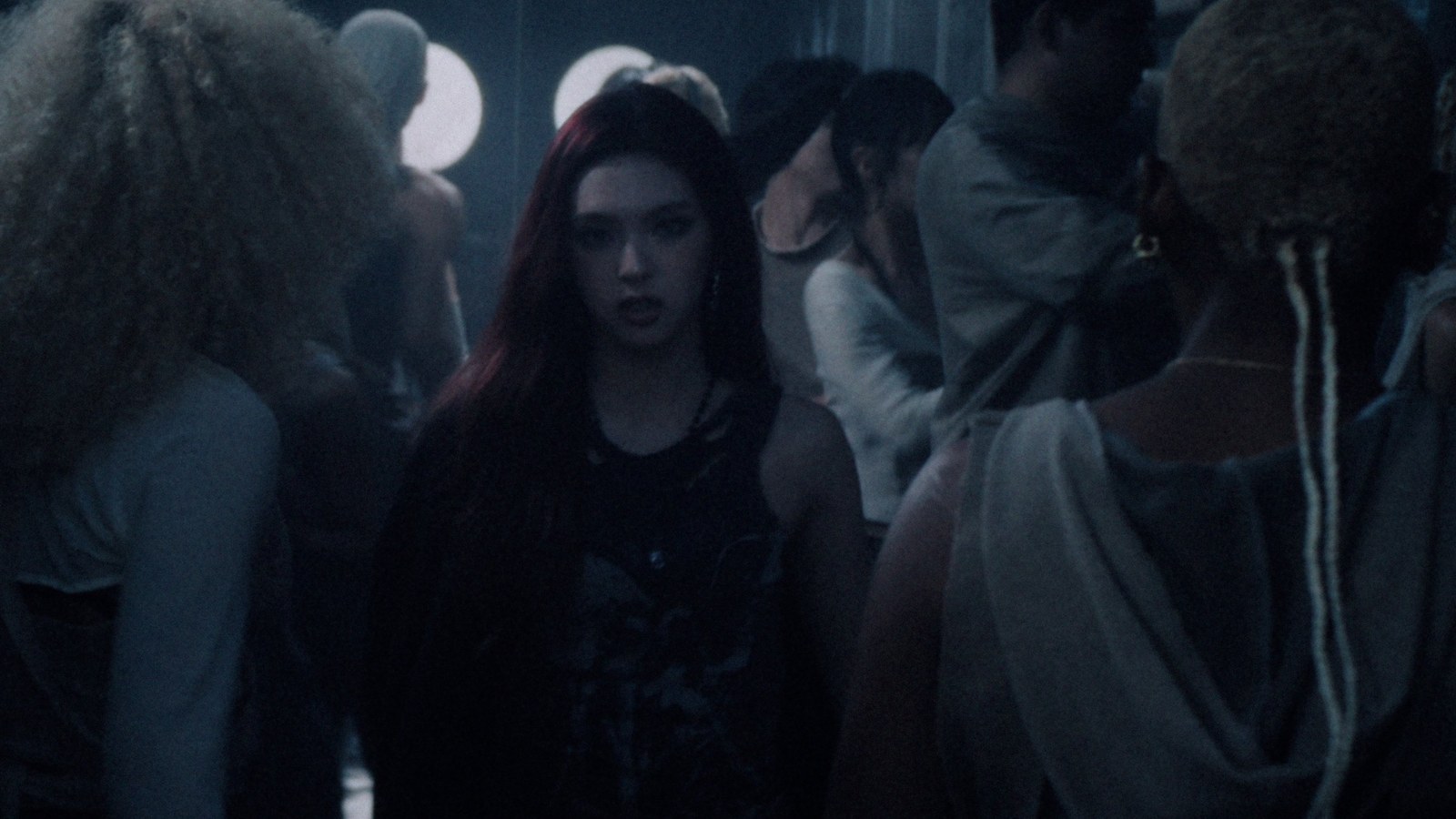 Member BABYMONSTER berambut merah berjalan di tengah kerumunan dalam MV DESERVE