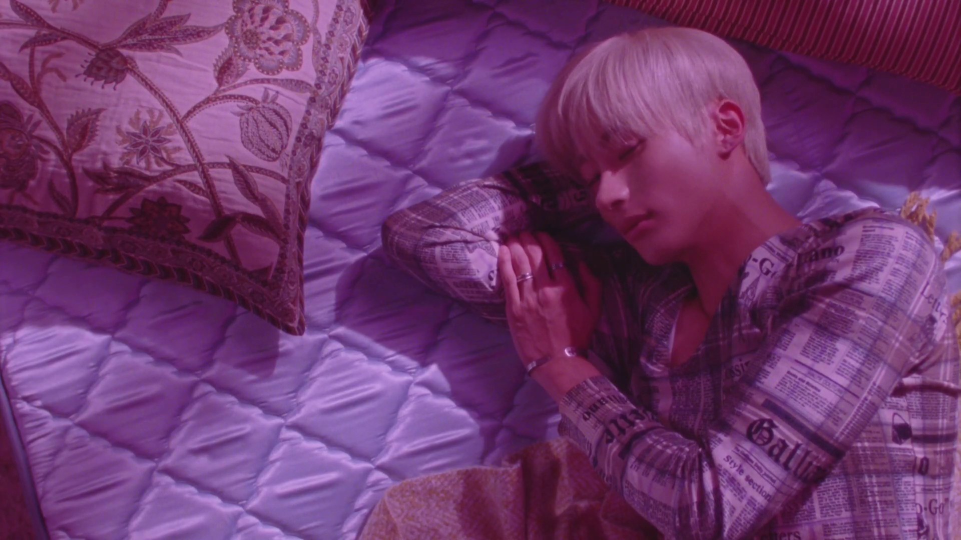 Member RIIZE berambut pirang tertidur di atas kasur dalam cahaya ungu pada teaser MV Insomnia